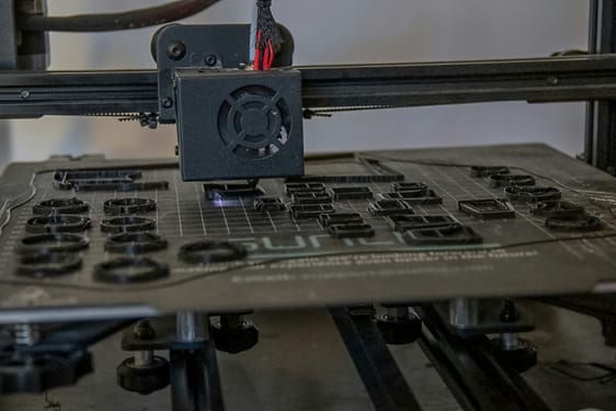 A 3D printer nozzle printing several small black circular parts on the build plate. It illustrates the production of custom components and short runs.