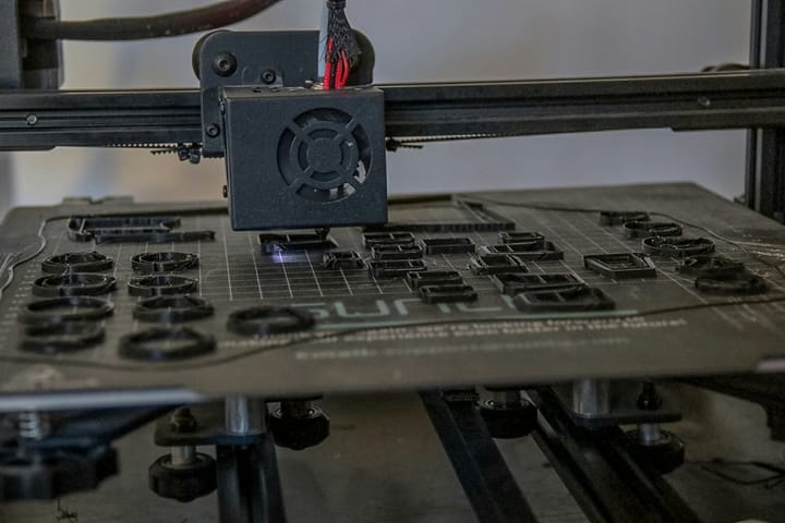 A 3D printer nozzle printing several small black circular parts on the build plate. It illustrates the production of custom components and short runs.