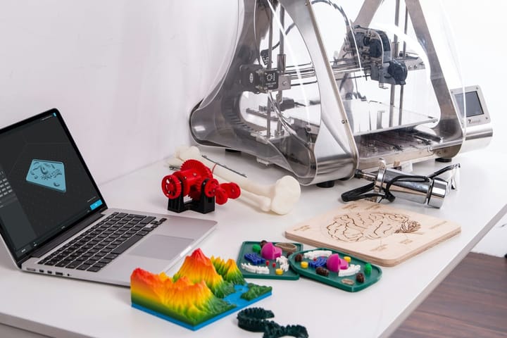 A desk with a laptop, a 3D printer beside it, and various 3D printed teaching models: a topographic map, a model of a human cell, and a gear with a bone. It illustrates the use of 3D printing in teaching.