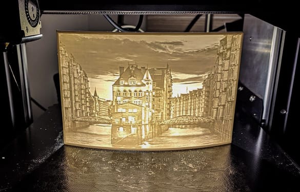 A 3D printer producing a pale, translucent image of a city with a river and buildings. The piece is a classic lithophane — a 3D printed photograph that glows when lit from behind, and an ideal personalised gift.