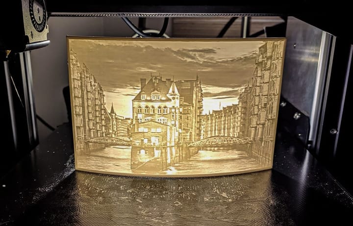 A 3D printer producing a pale, translucent image of a city with a river and buildings. The piece is a classic lithophane — a 3D printed photograph that glows when lit from behind, and an ideal personalised gift.