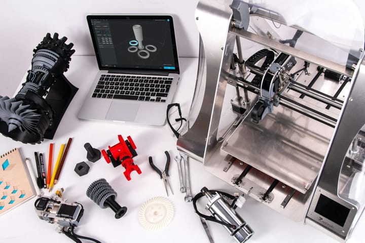 A workbench with an open laptop showing a 3D model and a 3D printer beside it. 3D printed prototypes are laid out around them — part of a jet engine, some gears — along with engineering tools. It illustrates the rapid prototyping process.